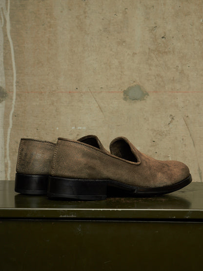 A pair of distressed brown leather slip-on shoes with black soles, displayed on a dark green metal surface against a textured beige concrete wall.
