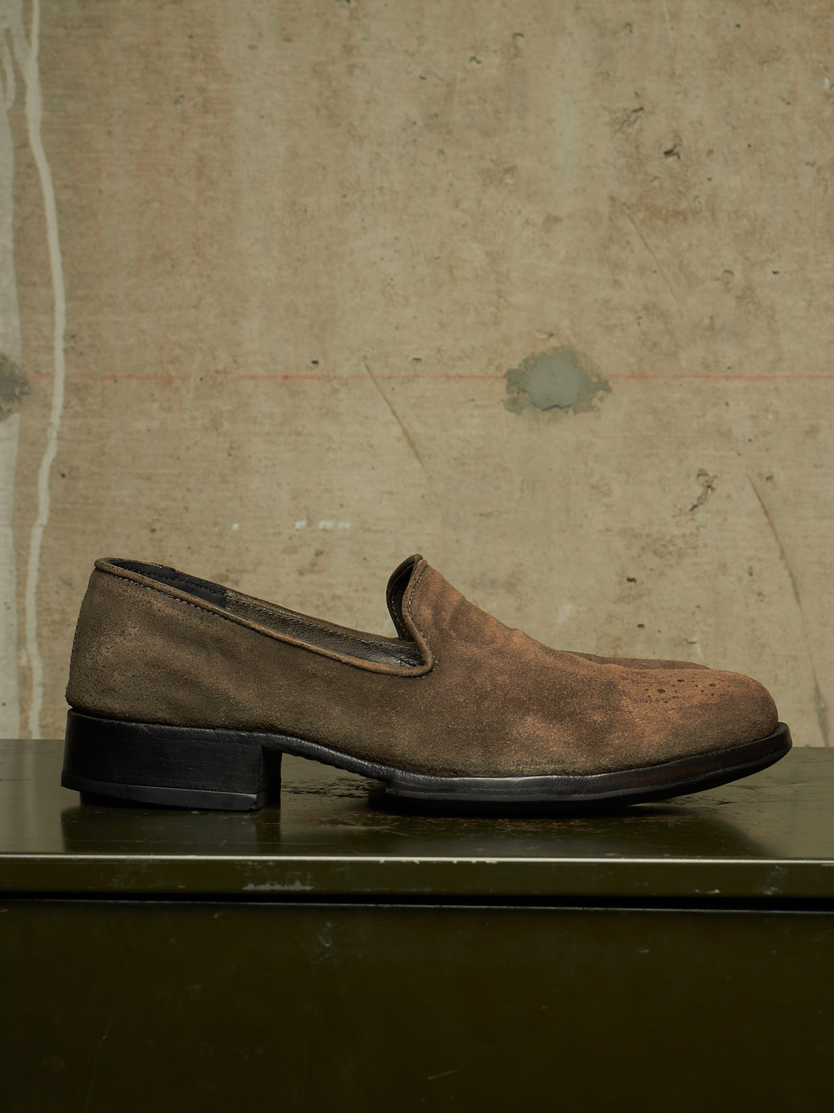 A single brown suede slip-on loafer with a low black block heel, resting on a dark green surface against a textured beige concrete wall.