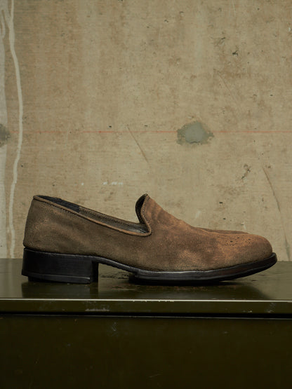 A single brown suede slip-on loafer with a low black block heel, resting on a dark green surface against a textured beige concrete wall.