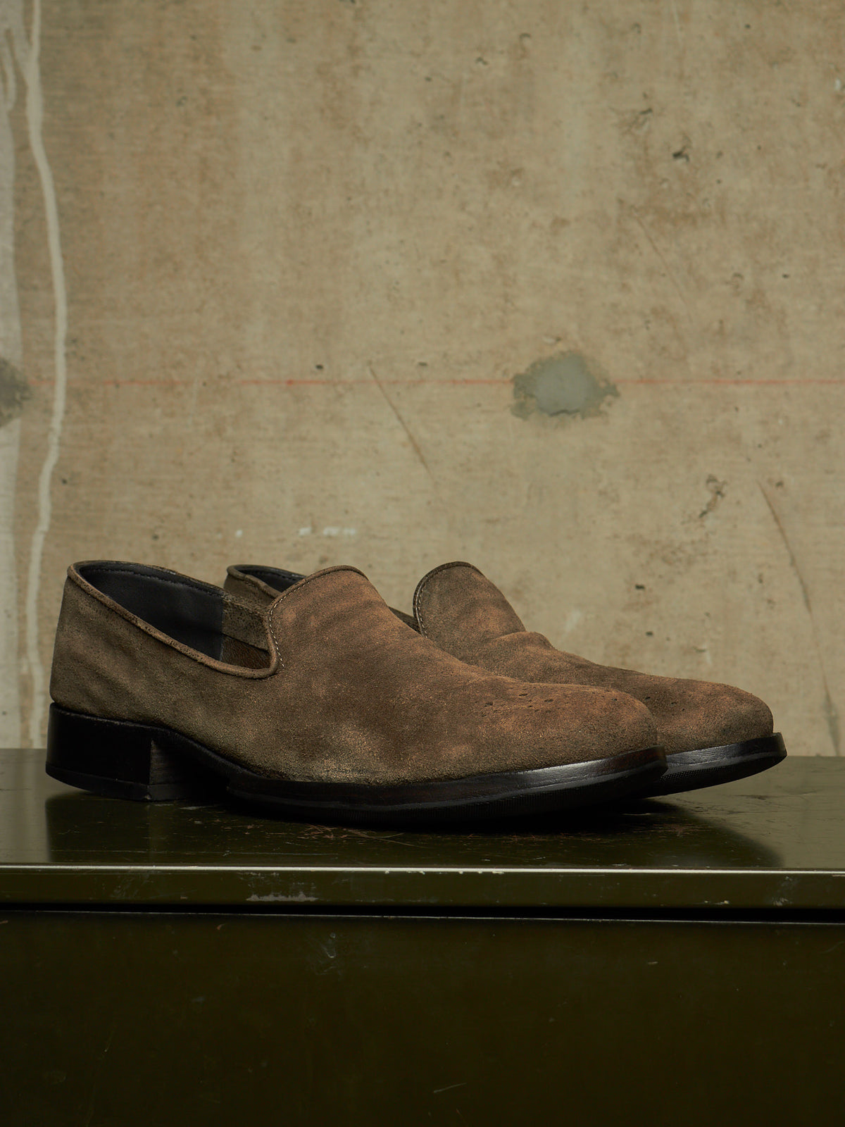 A pair of men's brown suede slip-on loafers with black soles, displayed on a dark green surface against a textured concrete wall.