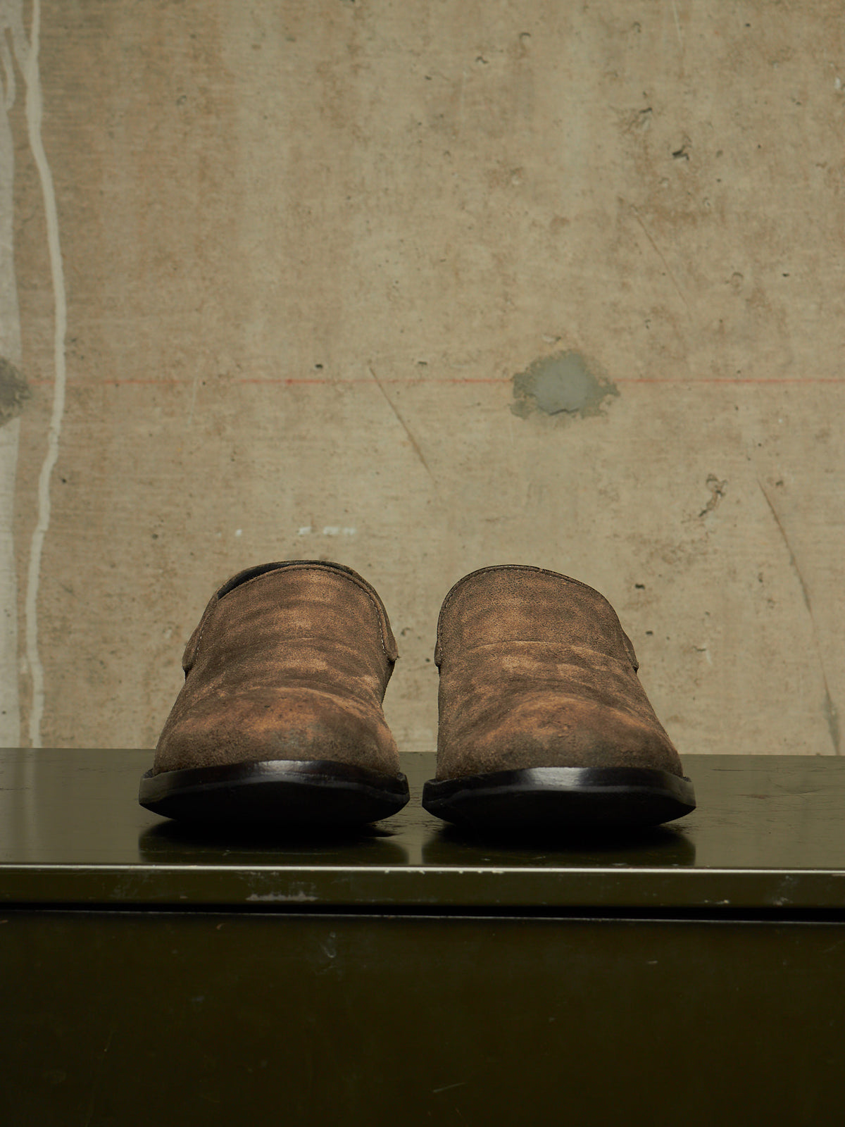 A pair of distressed brown leather slip-on shoes with black soles, displayed from the back on a dark green shelf against a textured beige wall.