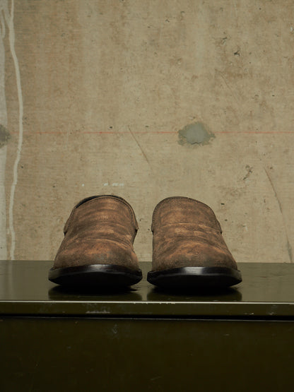A pair of distressed brown leather slip-on shoes with black soles, displayed from the back on a dark green shelf against a textured beige wall.