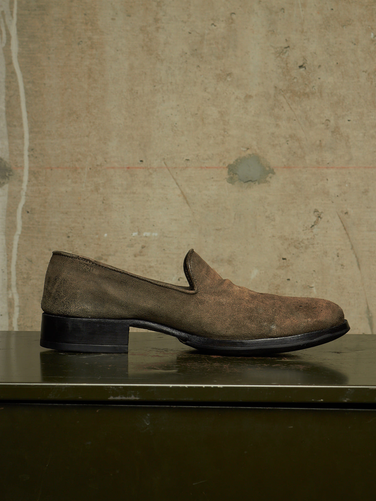 A single brown suede slip-on loafer with a low black block heel, resting on a dark reflective surface against a textured beige concrete wall.