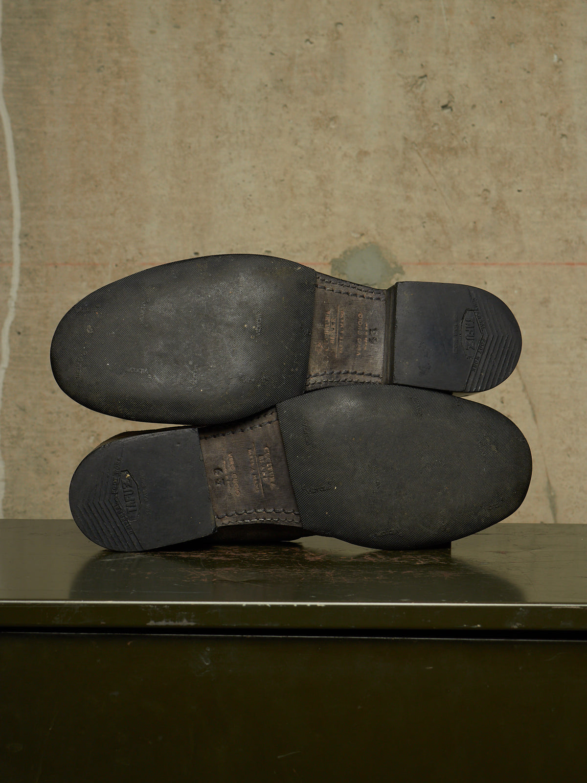 The soles of a pair of dark brown leather boots are shown from underneath, resting on a dark green metal surface against a textured beige wall.
