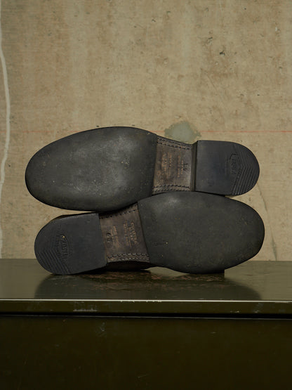 The soles of a pair of dark brown leather boots are shown from underneath, resting on a dark green metal surface against a textured beige wall.