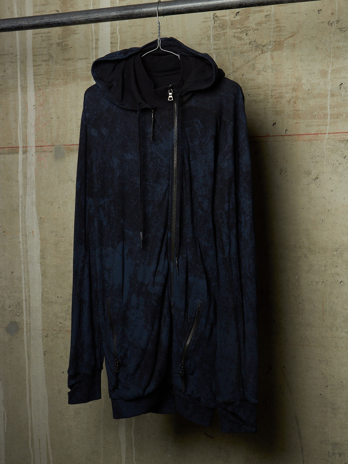 A dark navy hooded zip-up jacket with a mottled, textured pattern, hanging on a black hanger from a metal rail against a concrete wall.
