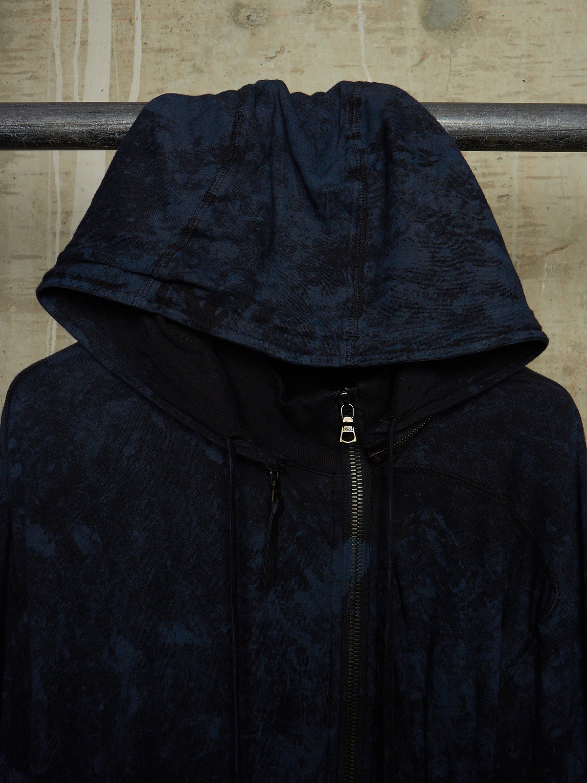 A dark navy hooded jacket with a textured, marbled pattern, hanging on a metal rack against a textured beige wall.