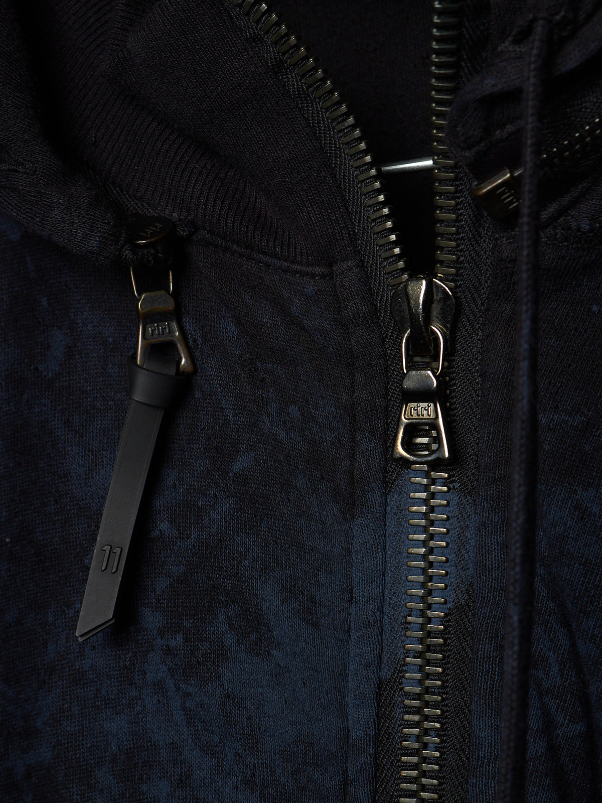 Close-up of a dark navy hooded jacket with a black zipper and a black pull tab featuring a small white logo, set against a dark textured fabric background.