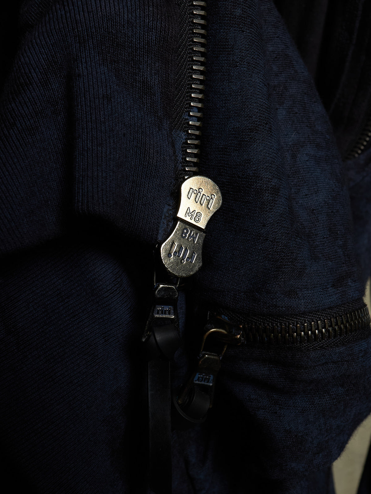 Close-up of a dark navy textured zippered garment with a silver-tone metal zipper pull engraved with 'mini MB' and 'BAH JUD'.