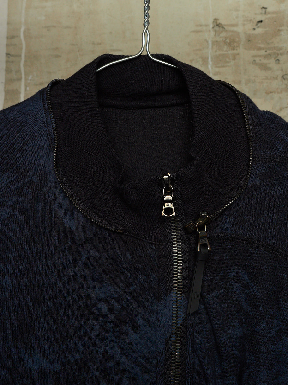 A close-up of a dark navy blue textured zip-up jacket hanging on a metal hanger against a light beige stone wall.
