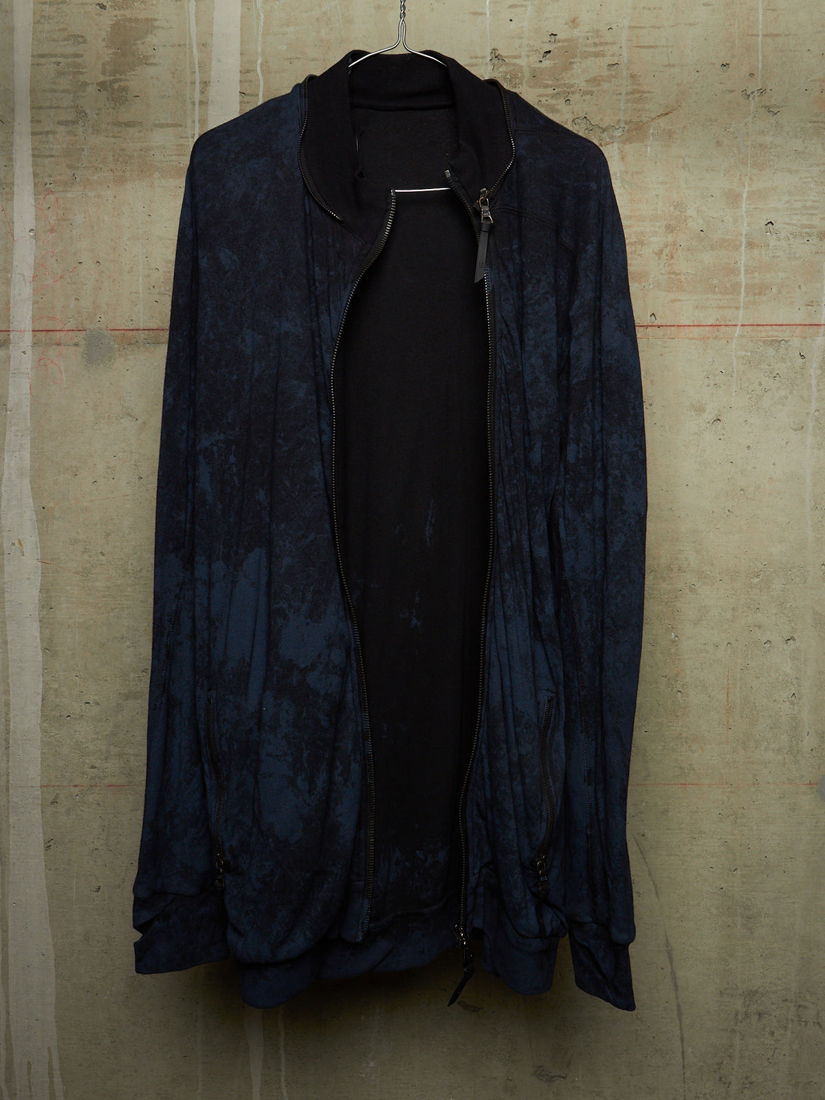 A dark navy blue lightweight jacket with a subtle marbled pattern, hanging open over a black top against a textured concrete wall.