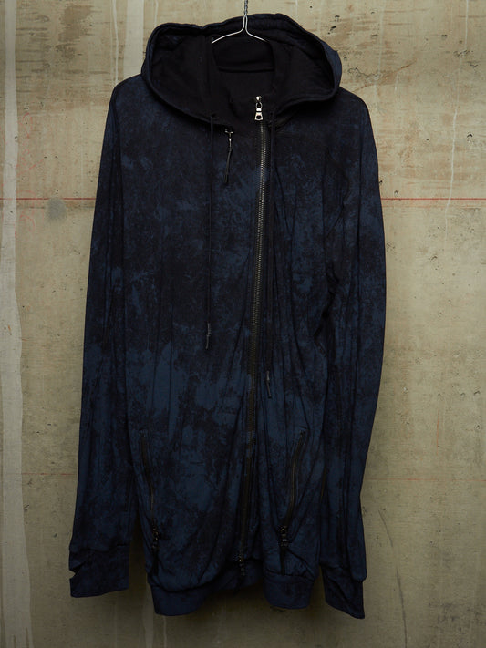 A dark navy blue hooded zip-up jacket with a mottled, tie-dye-like pattern, hanging against a textured concrete wall.