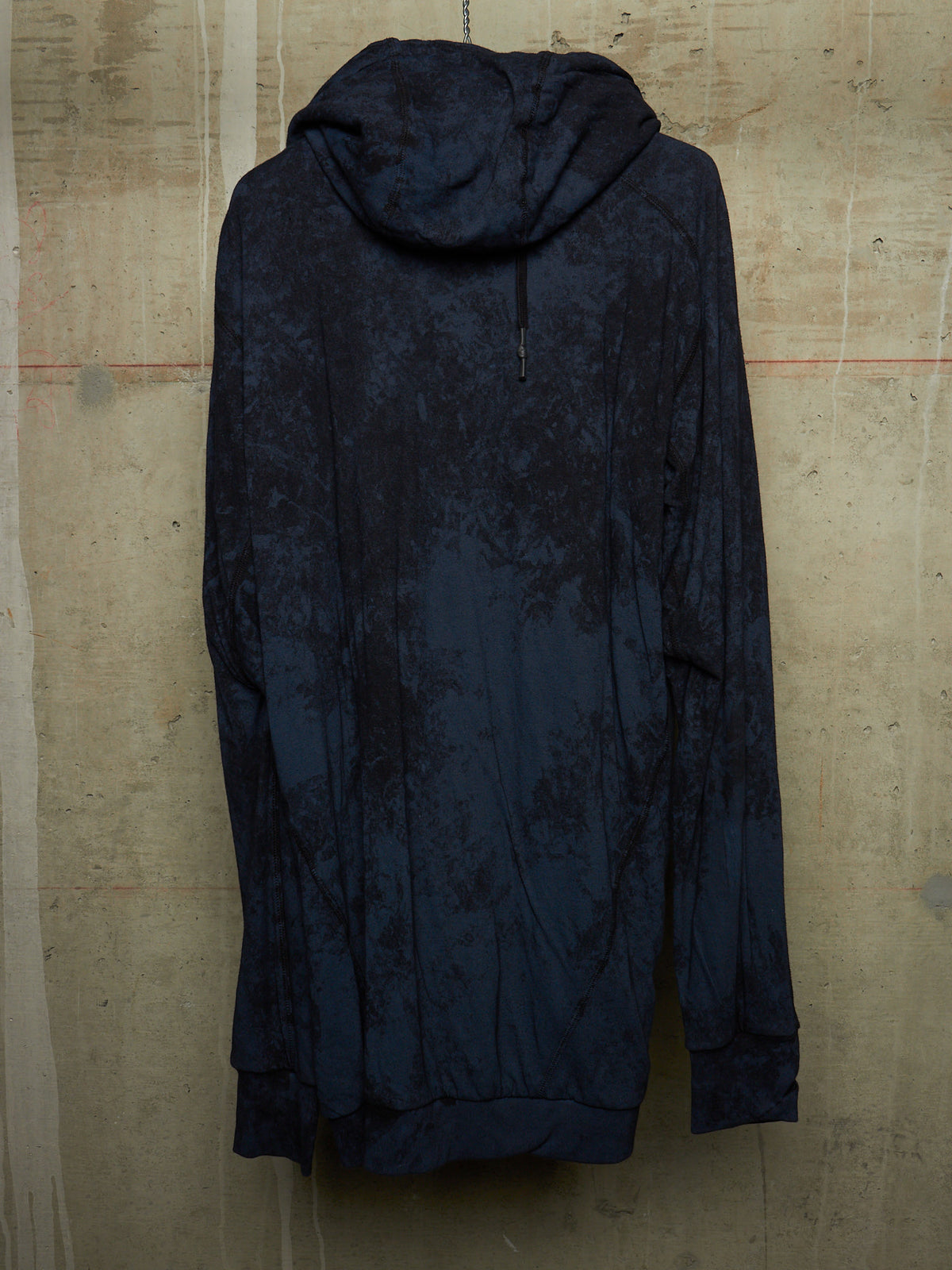A dark navy hooded sweatshirt with a mottled, tie-dye pattern, hanging against a textured concrete wall.