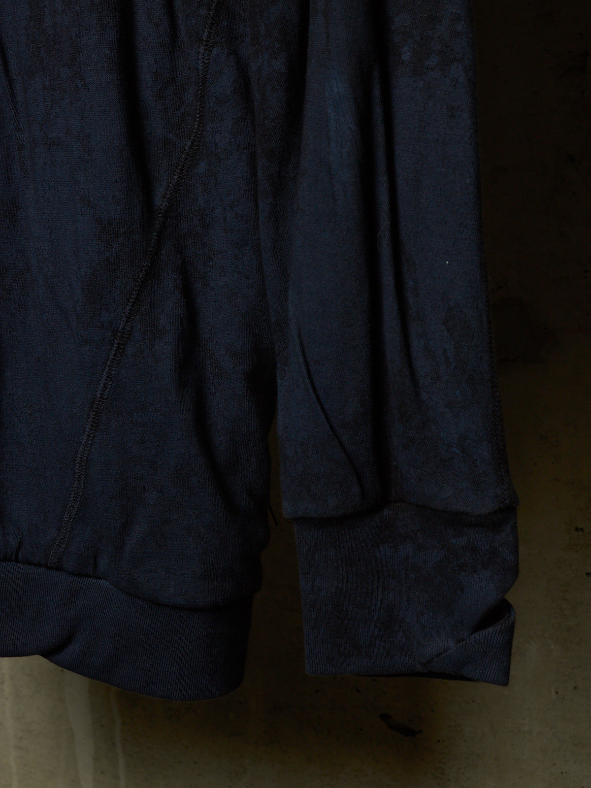 Close-up of a dark navy blue textured fabric garment, showing the cuff of a sleeve and the side seam against a dim, warm-toned background.