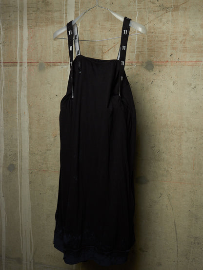 A black sleeveless dress with wide shoulder straps featuring a white and black pattern, hanging on a white wire hanger against a textured concrete wall.