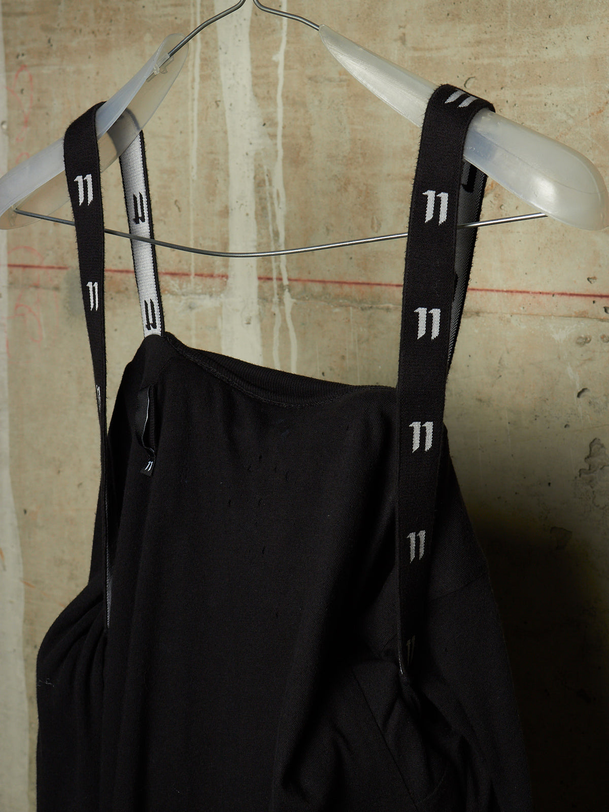 A black sleeveless top or dress with wide shoulder straps featuring a repeating white '11' pattern, hanging on a white hanger against a textured beige wall.
