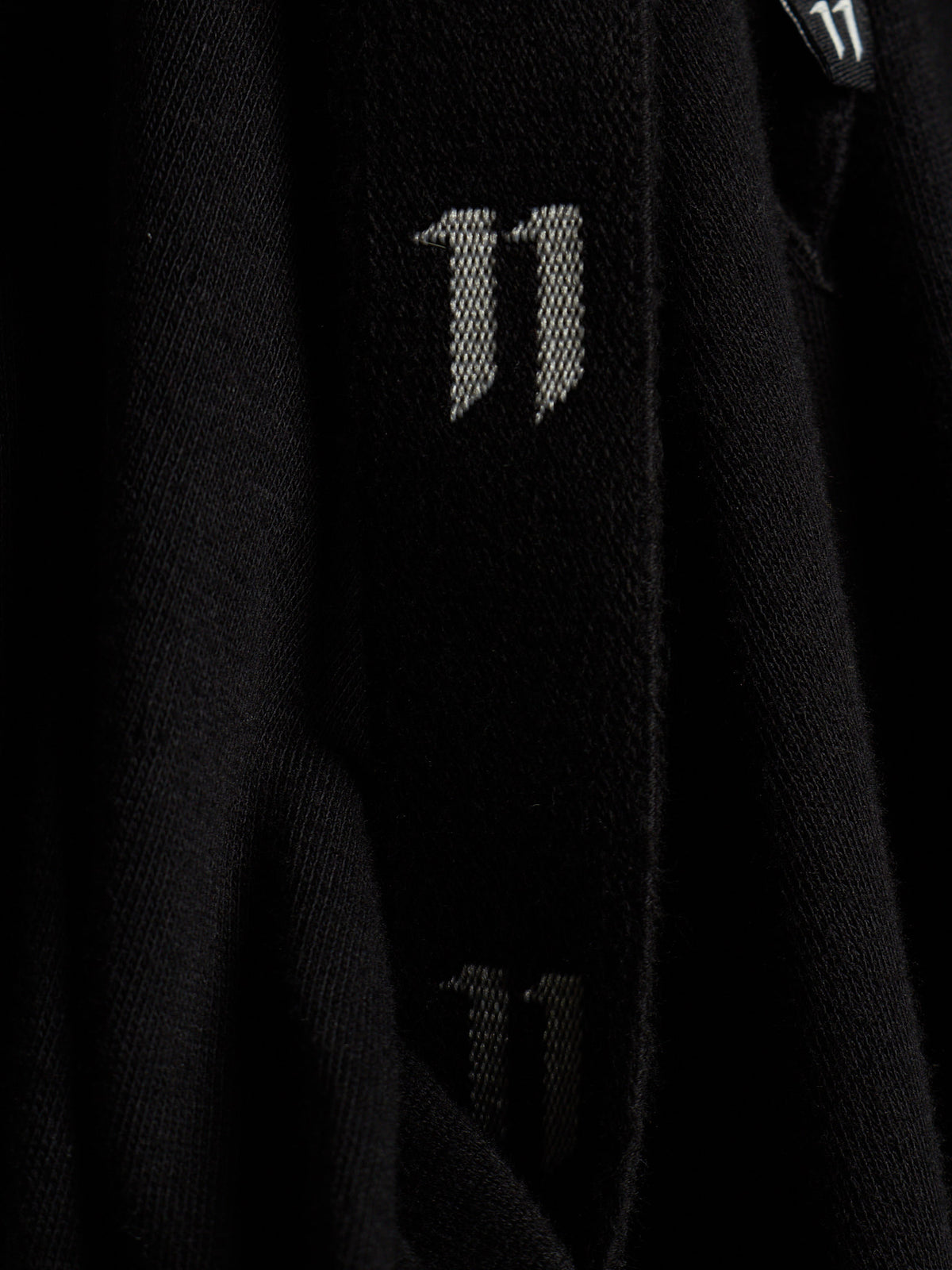 Close-up of a black knit garment featuring a large white stylized '11' logo on the sleeve, with a smaller matching logo visible near the collar.