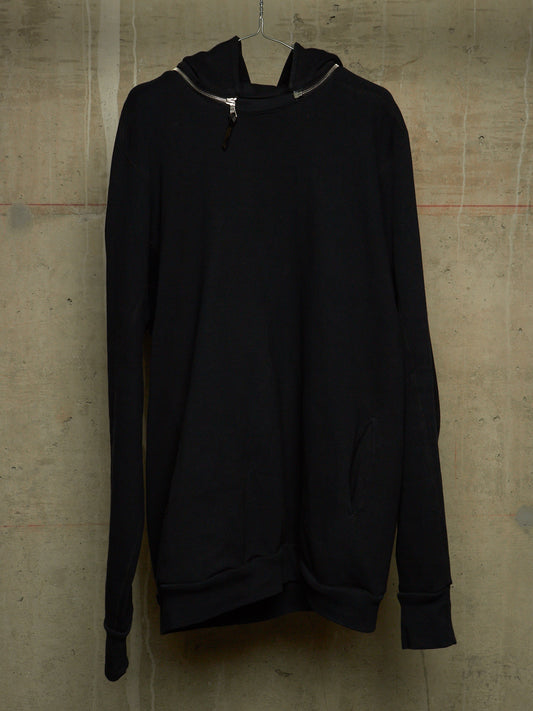 A black long-sleeve pullover hoodie with a half-zip neckline, hanging against a textured concrete wall.