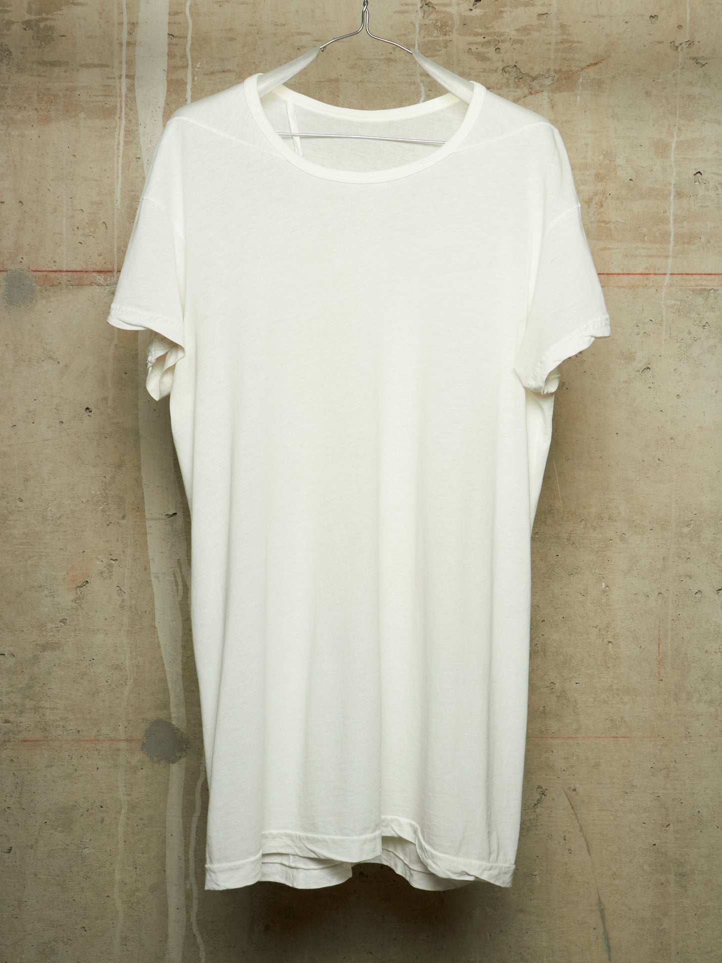 A plain white short-sleeve t-shirt hanging on a metal hanger against a textured concrete wall.