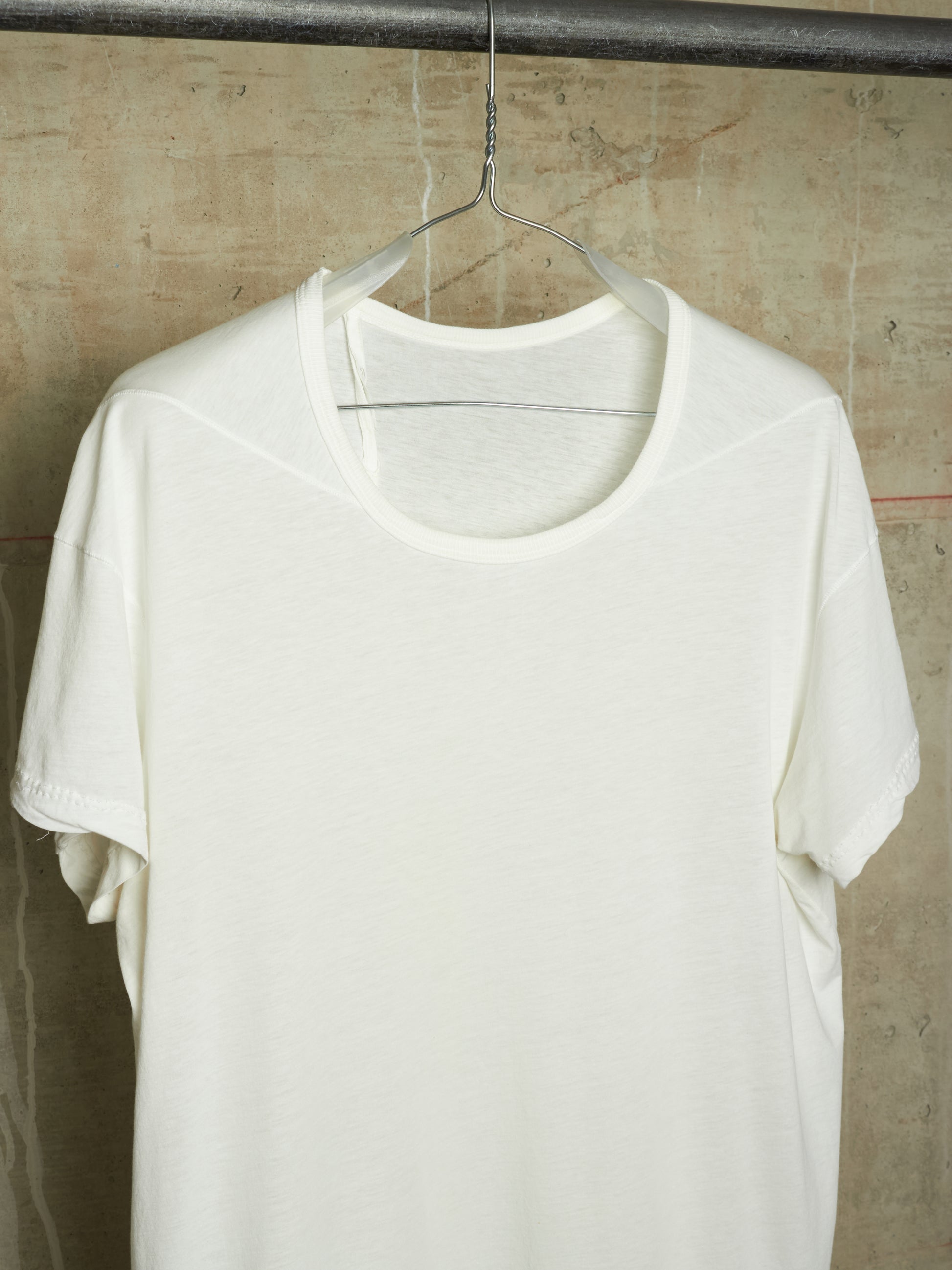 A white short-sleeve crewneck t-shirt hanging on a clear plastic hanger against a textured concrete wall.