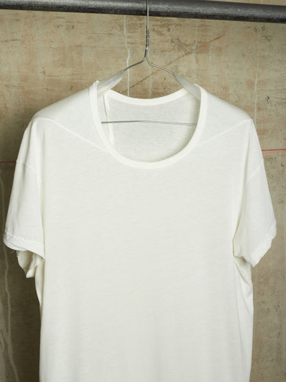 A white short-sleeve crewneck t-shirt hanging on a clear plastic hanger against a textured concrete wall.