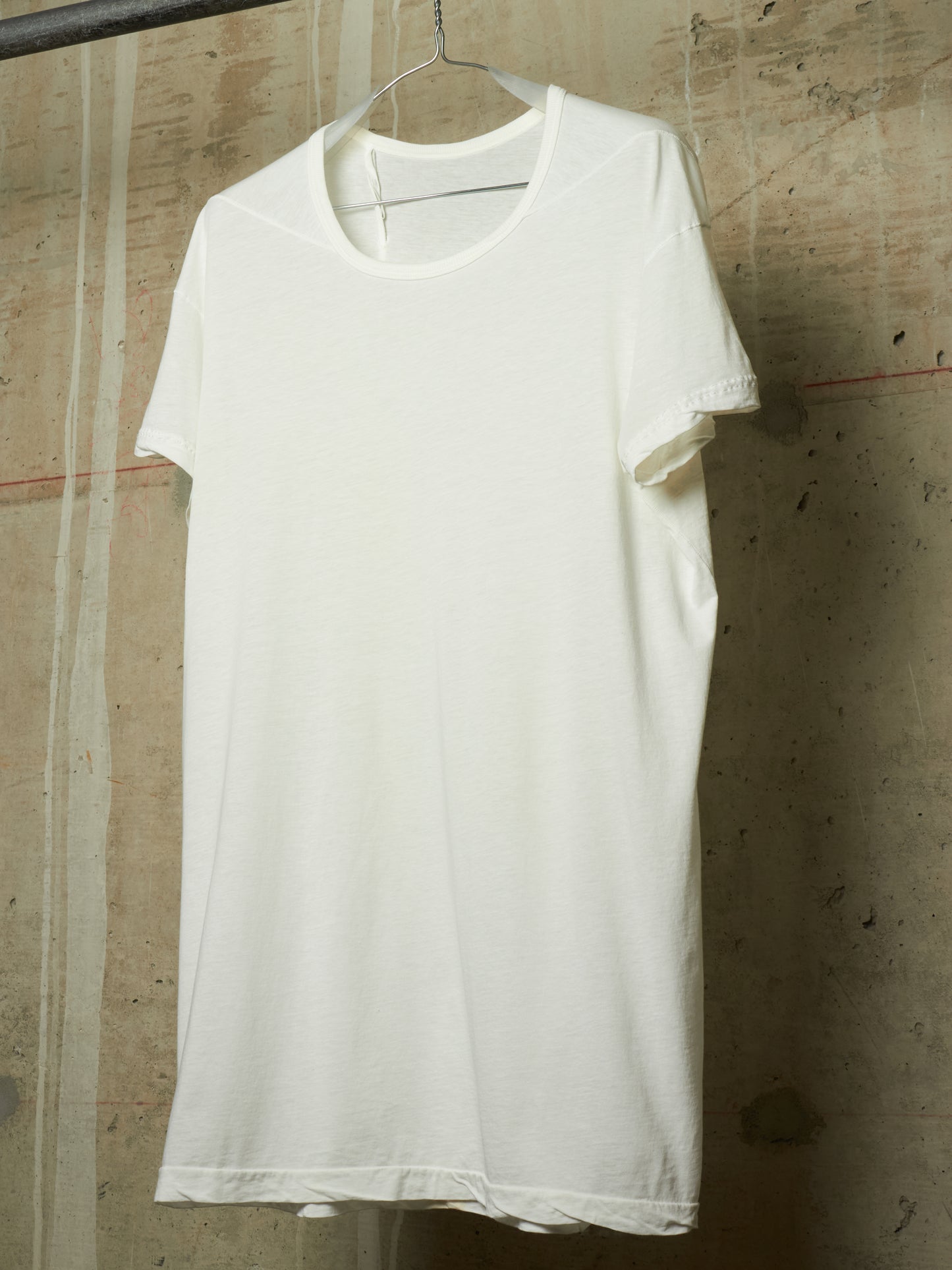 A plain white short-sleeve crewneck T-shirt hanging on a metal hanger against a textured concrete wall.