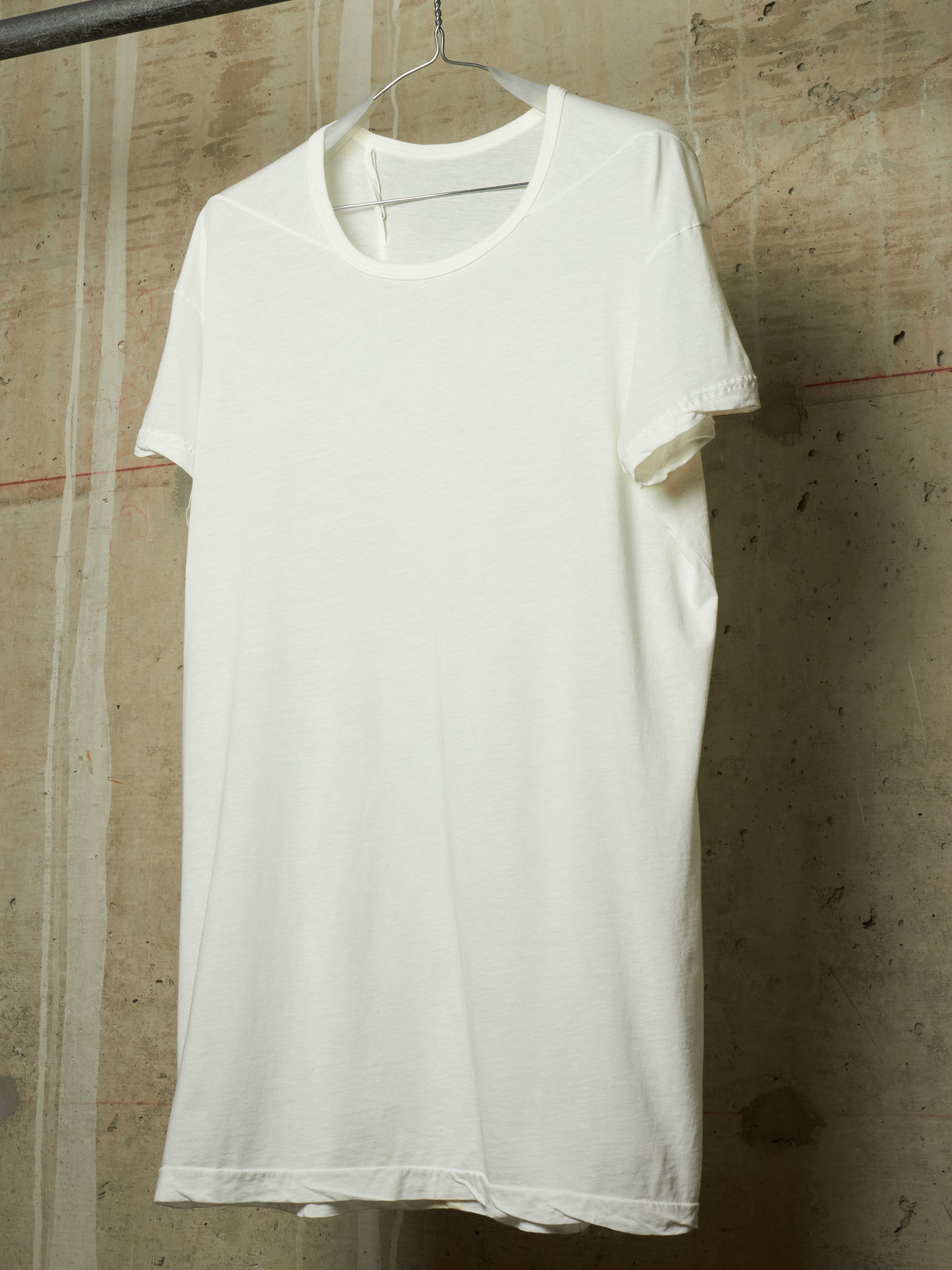 A plain white short-sleeve crewneck T-shirt hanging on a metal hanger against a textured concrete wall.