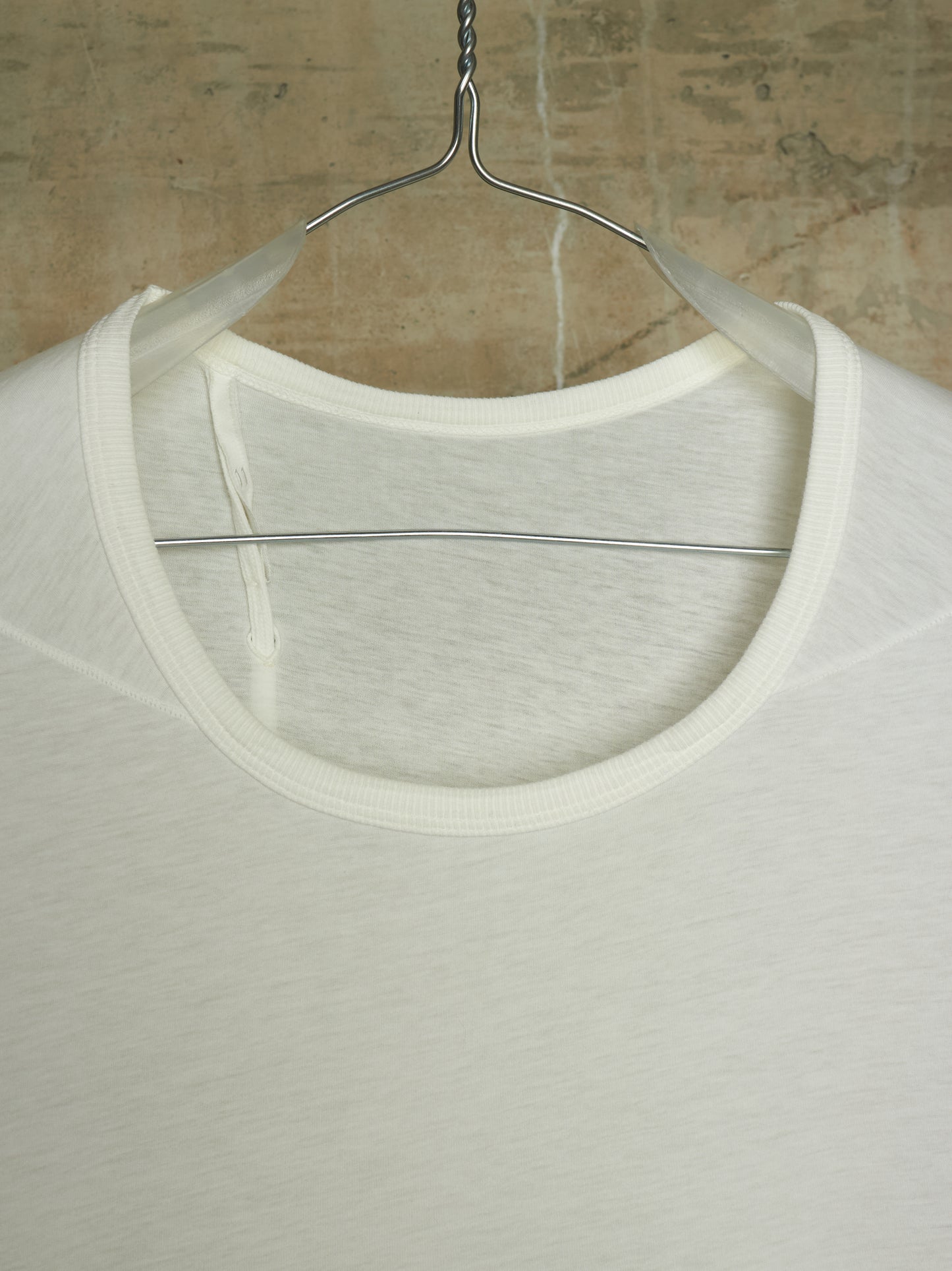 A close-up of a white crew-neck t-shirt hanging on a metal hanger against a textured concrete wall.