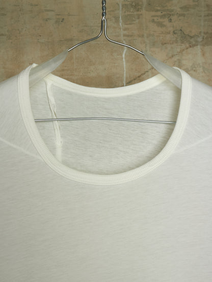 A close-up of a white crew-neck t-shirt hanging on a metal hanger against a textured concrete wall.