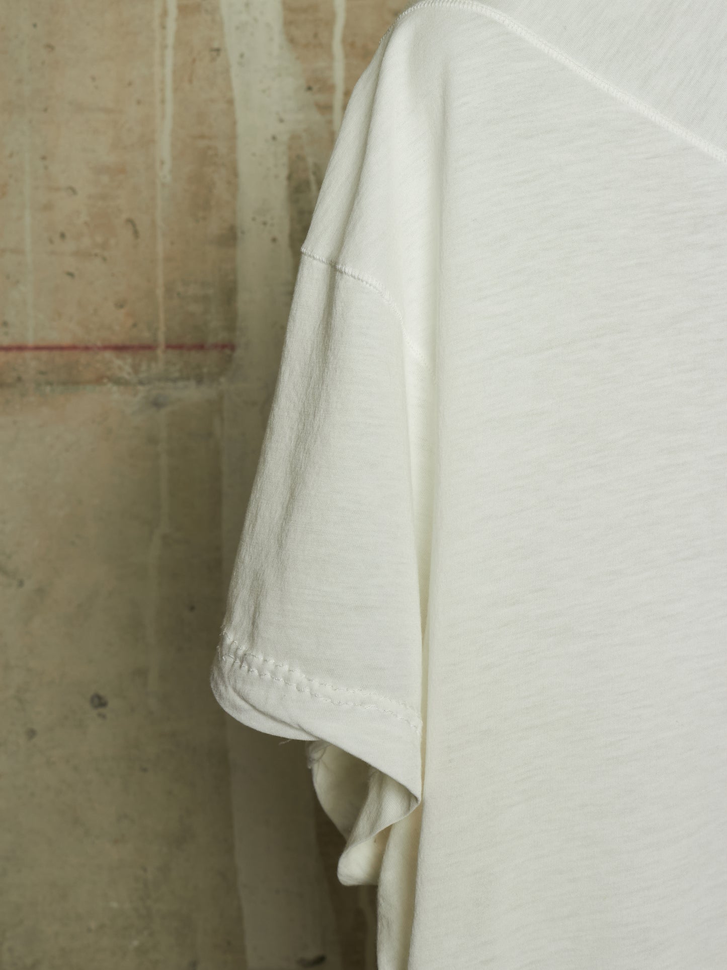 A close-up of an off-white cotton t-shirt sleeve and shoulder, showing the texture of the fabric and simple hem stitching, set against a rustic concrete wall background.