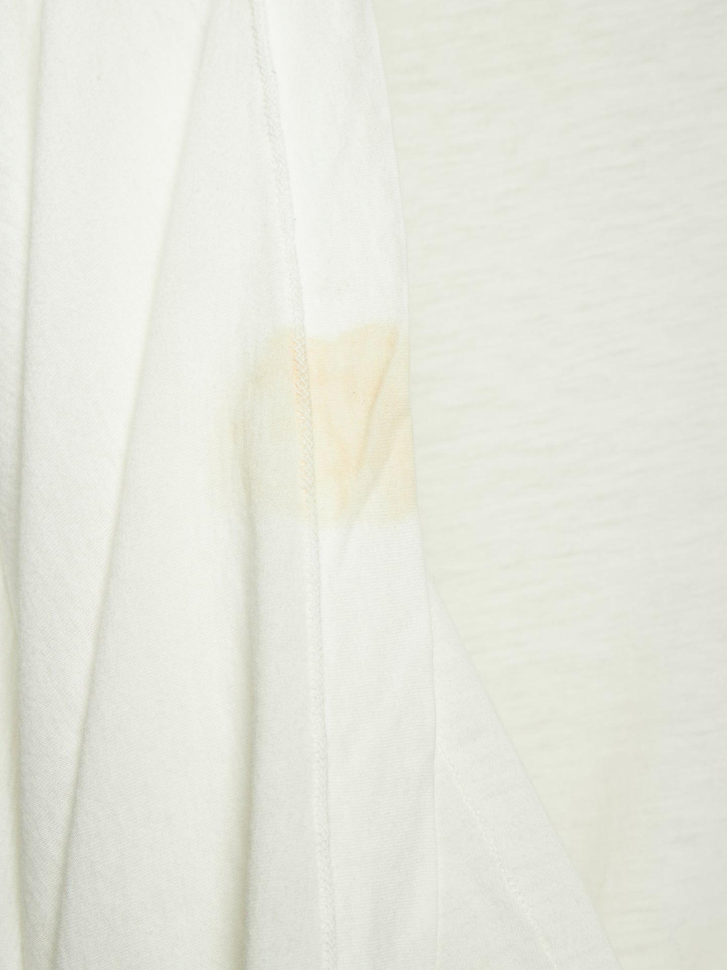 A close-up of a white textured fabric, likely a t-shirt or jersey, showing a light beige stain or discoloration on the left side seam.