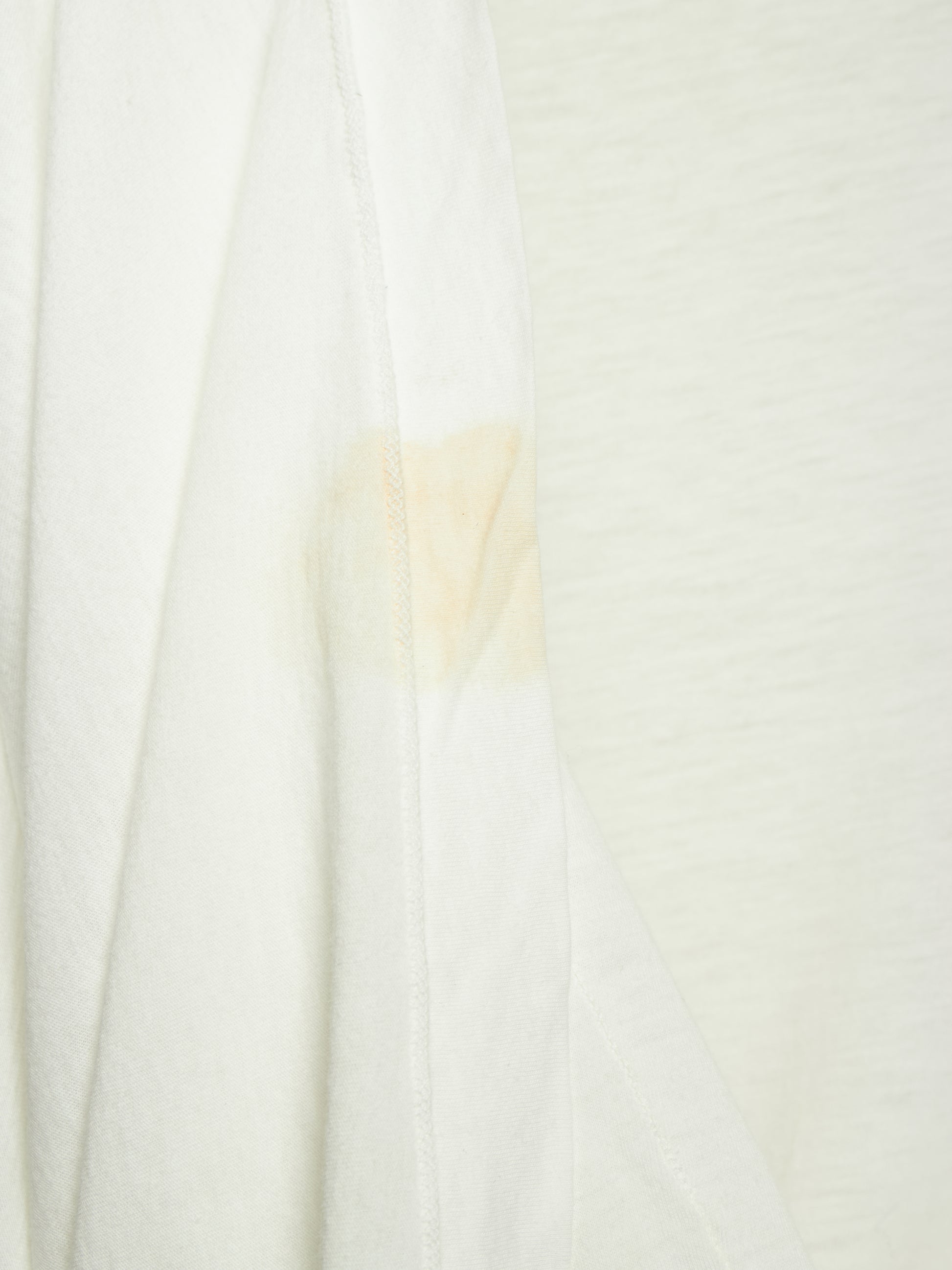 A close-up of a white textured fabric, likely a t-shirt or jersey, showing a light beige stain or discoloration on the left side seam.