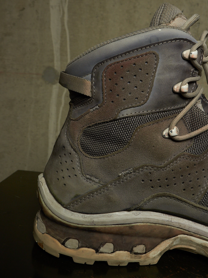 A close-up of a rugged gray hiking boot with suede and mesh panels, lace-up closure, and a thick sole, photographed against a dark textured background.
