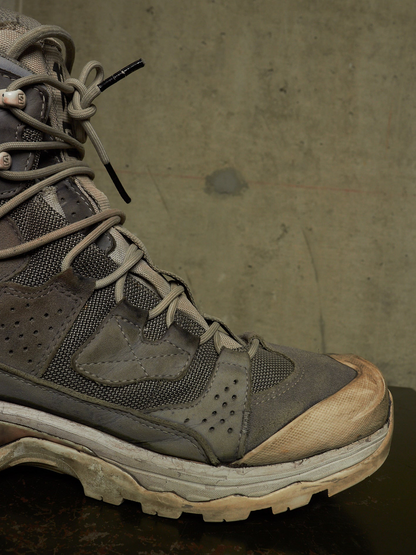 A close-up of a rugged gray and brown hiking boot with a thick off-white sole, resting on a dark surface against a textured concrete wall.