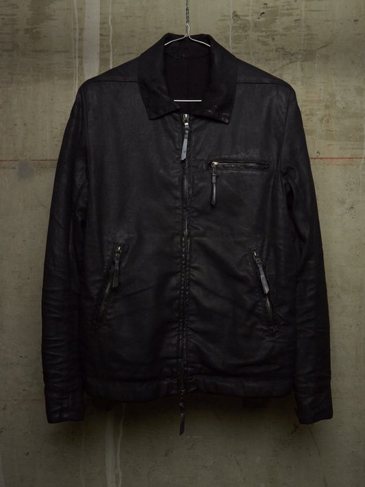 A black leather jacket hanging on a wire hanger against a textured concrete wall. The jacket features a zippered front, a zippered chest pocket, and zippered hand pockets.