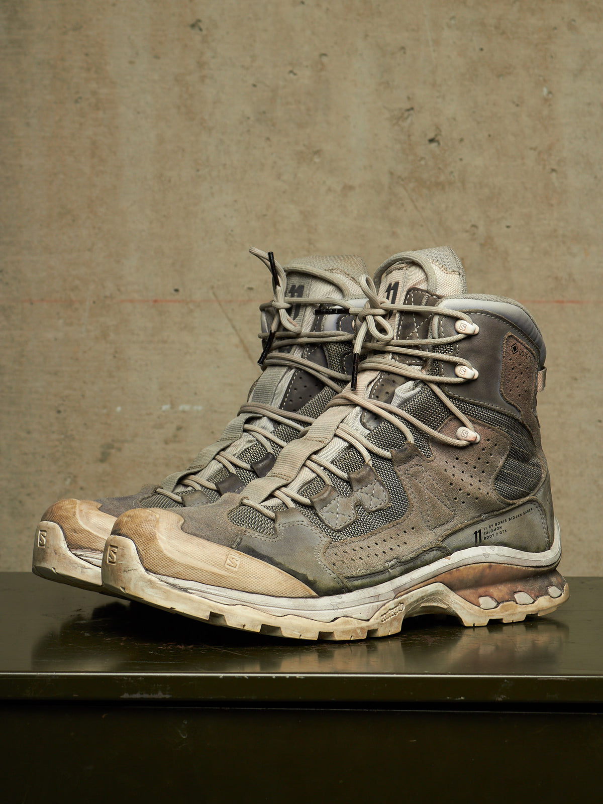 A pair of rugged lace-up hiking boots in distressed gray, brown, and beige synthetic materials, resting on a dark reflective surface against a textured concrete wall.