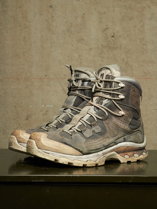 A pair of rugged lace-up hiking boots in distressed gray, brown, and beige synthetic materials, resting on a dark reflective surface against a textured concrete wall.
