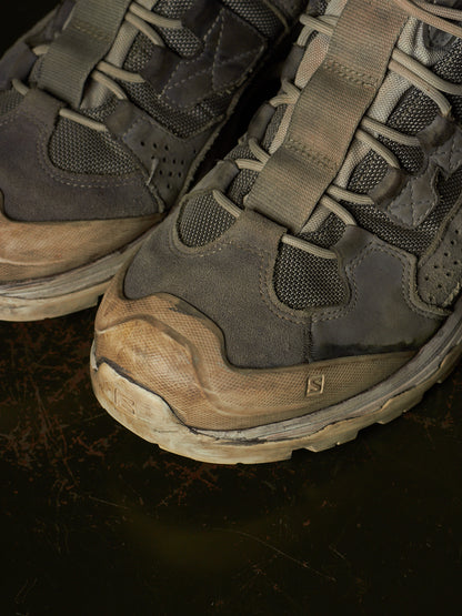 A close-up of a pair of rugged gray and brown hiking shoes with muddy, worn soles, resting on a dark textured surface.