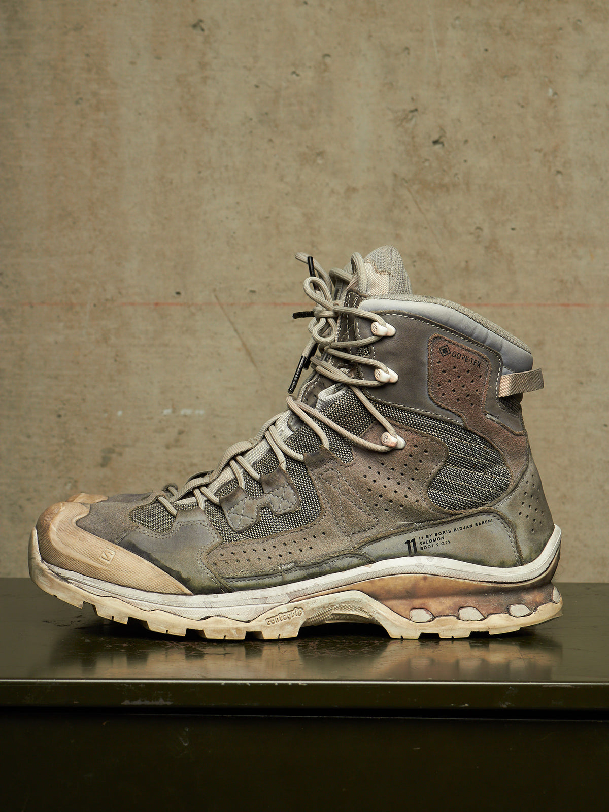 A single high-top hiking boot in distressed gray and brown synthetic materials, resting on a dark reflective surface against a textured concrete wall.
