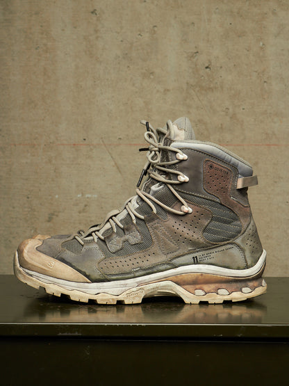 A single high-top hiking boot in distressed gray and brown synthetic materials, resting on a dark reflective surface against a textured concrete wall.