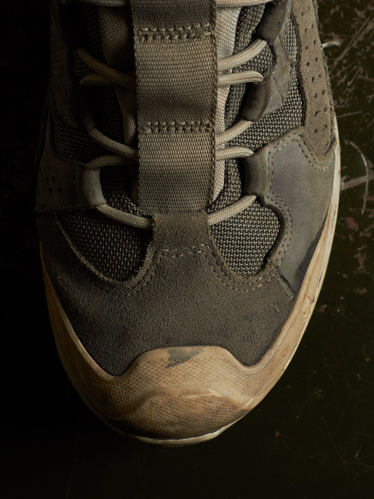 A close-up of a dark gray and brown suede and mesh sneaker with a thick off-white sole, shown against a dark background.