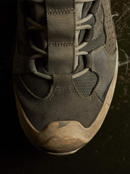 A close-up of a dark gray and brown suede and mesh sneaker with a thick off-white sole, shown against a dark background.