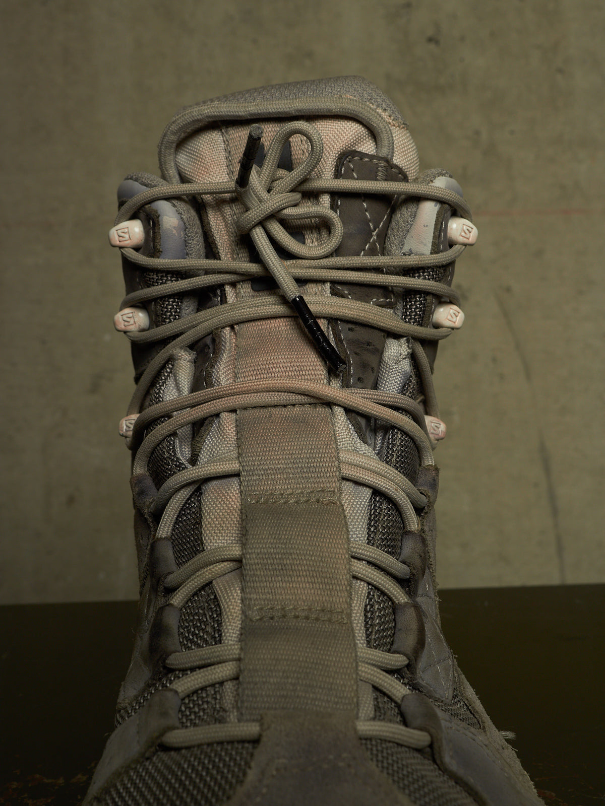 A close-up of a gray and brown tactical boot with thick laces threaded through metal eyelets, set against a dark textured background.