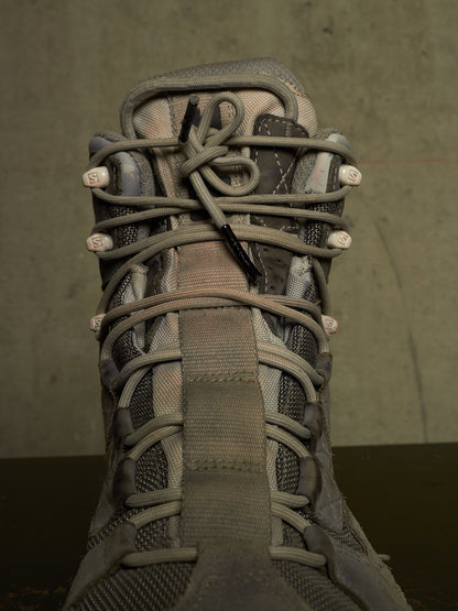 A close-up of a gray and brown tactical boot with thick laces threaded through metal eyelets, set against a dark textured background.