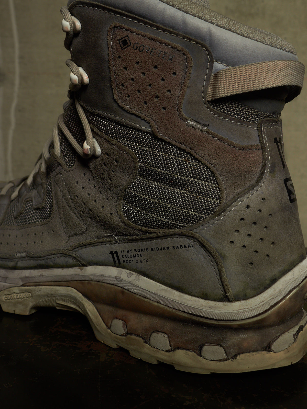 A close-up of a rugged hiking boot in dark brown and gray suede and mesh, featuring a pull tab, lace closure, and thick treaded sole.