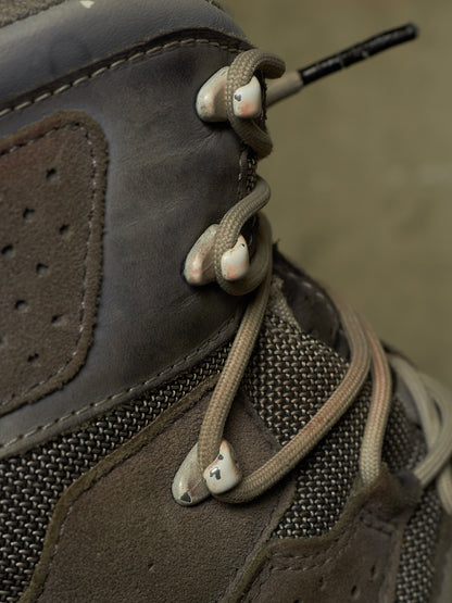 Close-up of a brown and gray high-top lace-up boot with beige laces threaded through metal eyelets.