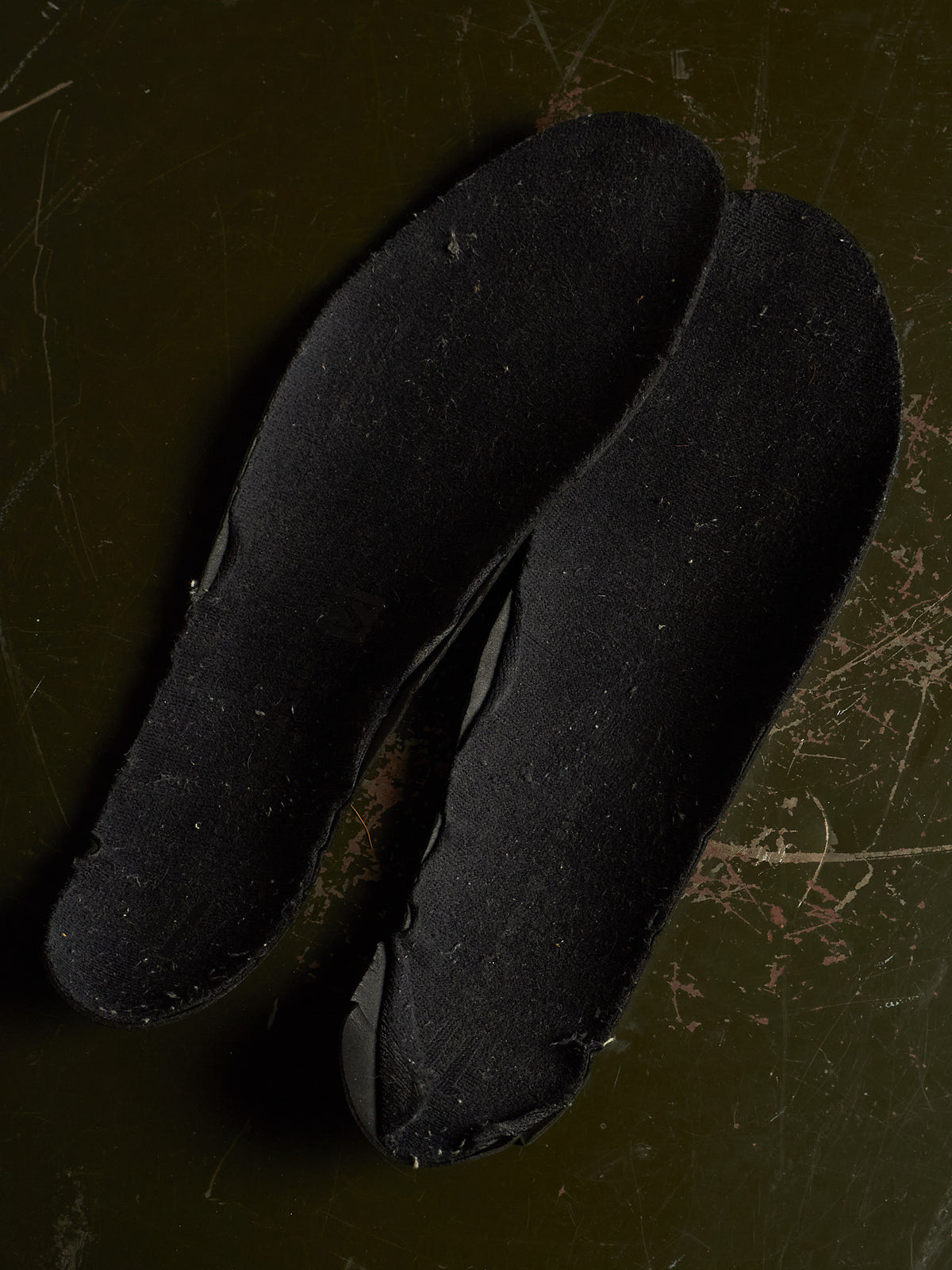 A pair of black fabric shoe insoles or footbeds laid flat on a dark olive-green textured surface.