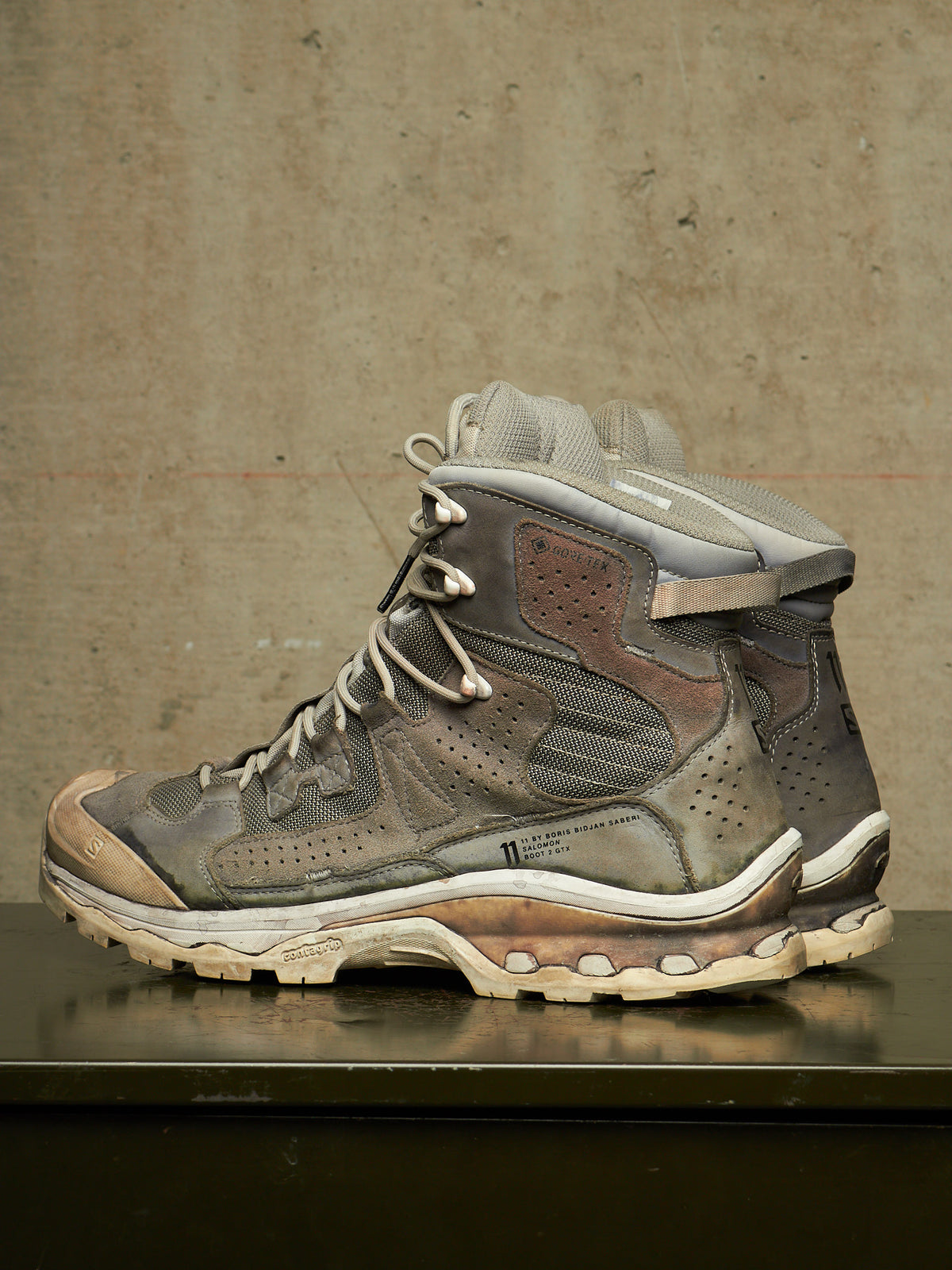 A single high-top athletic shoe in distressed gray, brown, and beige tones, photographed from the side against a textured concrete wall on a dark reflective surface.