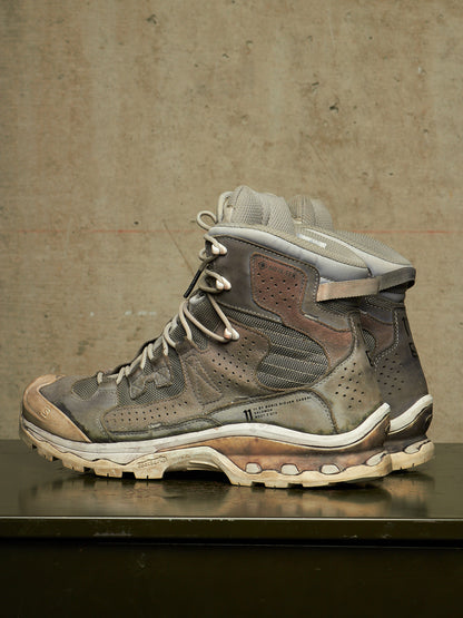 A single high-top athletic shoe in distressed gray, brown, and beige tones, photographed from the side against a textured concrete wall on a dark reflective surface.