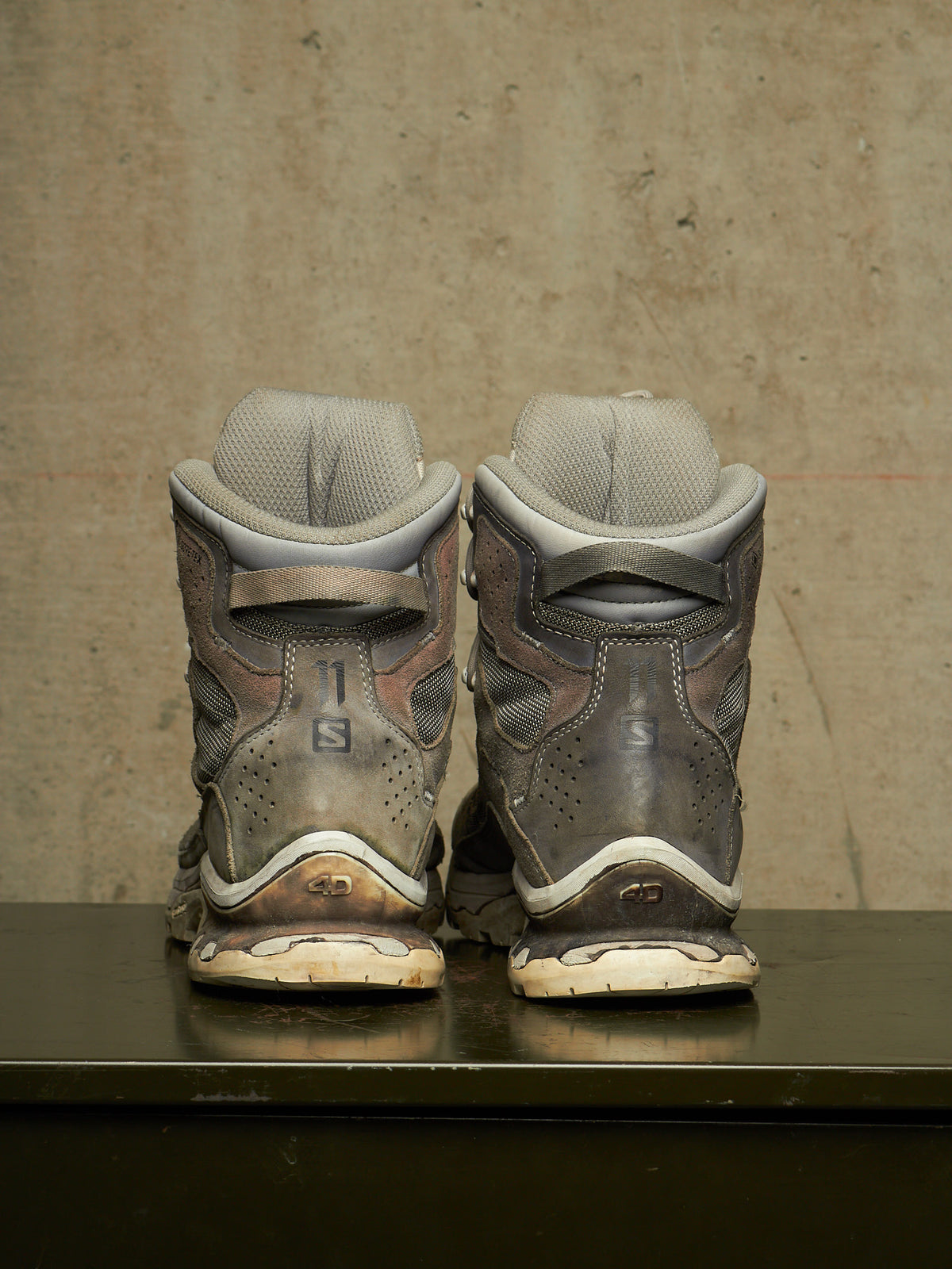 A pair of used Salomon snowboard boots shown from the back, positioned side-by-side on a dark reflective surface against a textured concrete wall.